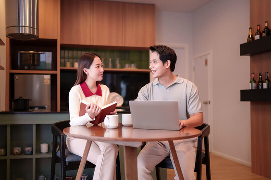 Asian Couple Working Together From Home Office on Laptop and Reading Book Joyful Conversation Collaboration at Kitchen Countertop
