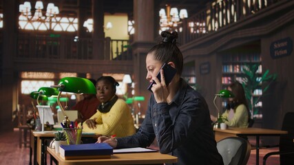 Noisy person disturbing people at the library by speaking very loud, being rude and answering a phone call in the study area. Disruptive annoying woman asked to keep silence. Camera A.