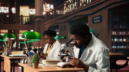 African American pupil studies microbiology in a campus library, examining cells and microorganisms under the microscope. Male student prepares for his biotechnology thesis. Camera A.