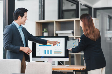 Business professionals discussing dashboard data analysis in modern office, man pointing at computer screen with charts while woman holds tablet, teamwork and collaboration concept