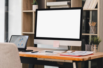 Modern workspace with large blank computer monitor, laptop displaying charts, smartphone, documents, and office supplies on wooden desk.