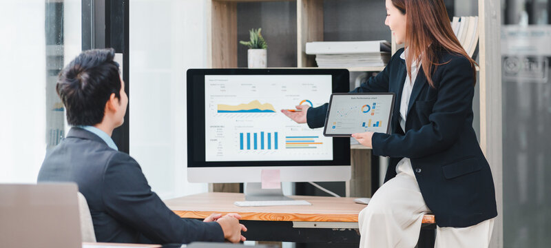 Businesswoman presenting dashboard data analysis to colleague using computer and tablet in modern office, discussing financial growth and performance metrics in professional setting