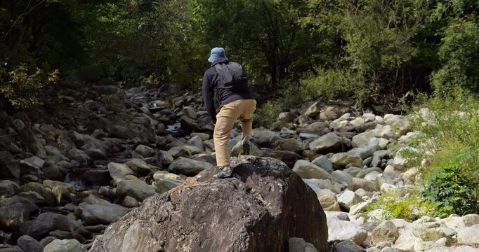 Tourist hiker man climbs onto large rock midstream and raises his arms in high spirits, surrounded by pristine mountain wilderness. moment captures sense of adventure and personal achievement