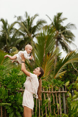 Mother lifting baby girl high into air with tropical foliage behind them.