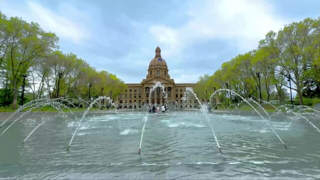 Edmonton, Alberta, Canada. May 22, 2025. The Alberta Legislature Building, framed by spring foliage and a fountain's spray, stands as a symbol of democracy under a cloudy sky.
