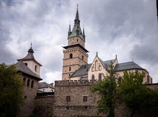 View of the fortified medieval Kremnica Castle with the Church of St. Catherine, surrounded by stone walls, towers, and greenery under a dramatic cloudy sky.
