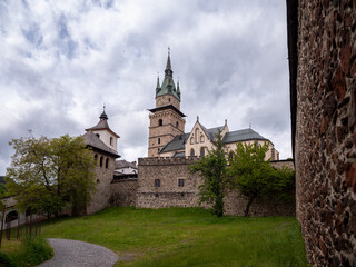 View of the fortified medieval Kremnica Castle with the Church of St. Catherine, surrounded by stone walls, towers, and greenery under a dramatic cloudy sky.
