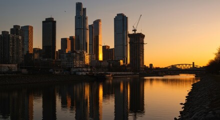 Fototapeta premium Cityscape Sunset Reflection - Stunning sunset view of a modern city skyline reflected in calm water, showcasing skyscrapers and a bridge