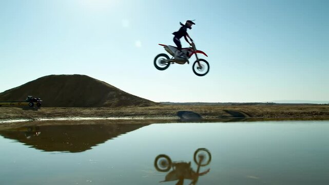 A motocross rider leaps off a dirt mound, casting a reflection in a nearby pond under a clear blue sky - moto stunt bike
