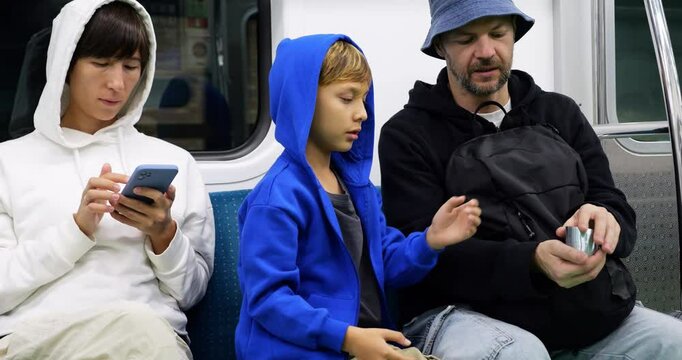 Father shows young son how to sway slinky from hand to hand while riding Seoul subway in evening, mother sit beside focused on smartphone. Train in motion, flashing outdoors visible through window.