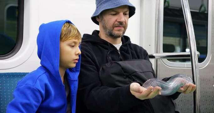 Father demonstrates how to play with metal spring toy during subway ride, while his son, once fascinated by rhythmic shimmer of slinky, now sits silently in thought, peering through coils of toy