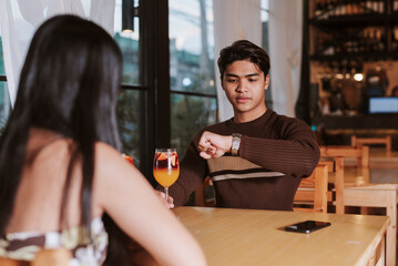 A young Southeast Asian man glances at his watch while seated across from his date, signaling...