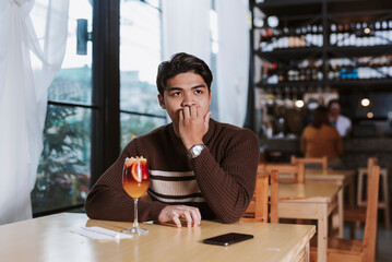 A young nervous Filipino man anxiously bites his nails while waiting alone at a restaurant, worried...