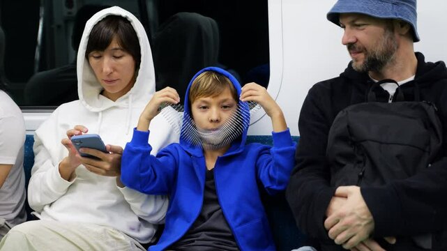 Family riding subway train, mother checking smartphone while father watching son playfully explore slinky toy during casual urban commute. Boy holds spring near his face, looking playfully