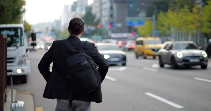 Professional businessman standing on roadside with backpack, waiting for taxi and looking ahead. Rear view waist-up slow motion shot with moderate daytime traffic on road. Selective focus on man