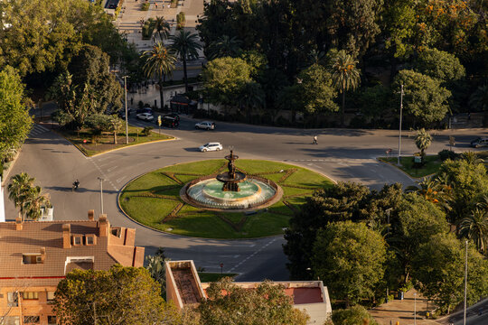 Aerial view of a circular fountain surrounded by lush greenery and palm trees, with vehicles navigating the roundabout in a vibrant urban setting showcasing city life