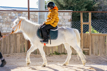 Boy in jacket and helmet ridding a white horse outdoors
