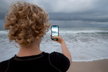 Teenage boy photographing a stormy sky at beach with phone