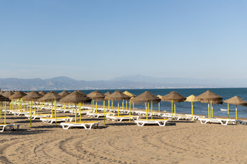 Beach scene featuring sun loungers and straw umbrellas on sandy shore with calm sea and distant mountains under clear blue sky