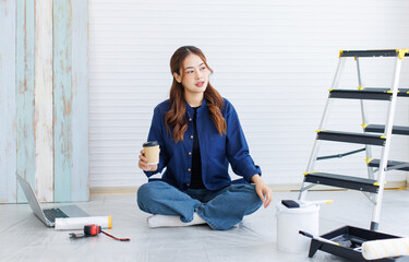 Asian young beautiful woman long curly hair wearing blue shirt jeans sit on floor holding coffee cup look for inspiration paint color walls around rollers brush ladder computer renovate room concept.