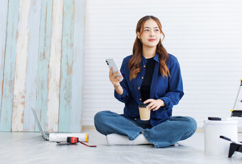 Asian young beautiful woman long curly hair wearing blue shirt jeans sit on floor using smartphone consult with friend short break after painting wall around rollers brush ladder renovate room concept