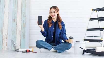 Asian young beautiful woman long curly hair wearing blue shirt jeans sit on floor using smartphone consult with friend short break after painting wall around rollers brush ladder renovate room concept