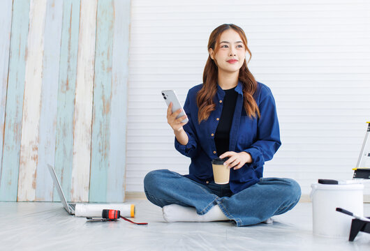 Asian young beautiful woman long curly hair wearing blue shirt jeans sit on floor using smartphone consult with friend short break after painting wall around rollers brush ladder renovate room concept - Powered by Adobe