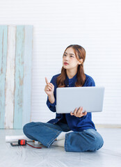 Asian young beautiful woman long curly hair wearing blue shirt jean sit on floor look at laptop computer learning before repairing room painting wall house or apartment rollers brush ladder background