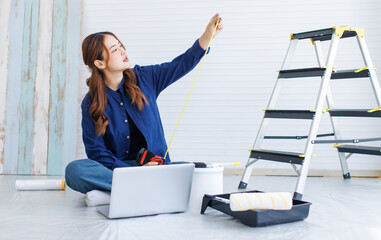 Asian young beautiful woman long curly hair wearing blue shirt jean sit on floor looking measure tape before repairing room painting wall empty house or apartment rollers brush ladder in background