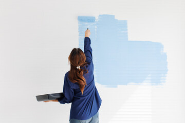 Asian young beautiful woman smiling cheerful long curly hair wearing blue shirt jeans sitting on ladder painting wall with roller brush light blue for new home repair renovation decoration concept.