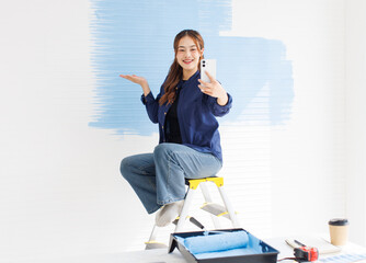 Asian young beautiful woman smiling cheerful long curly hair wearing blue shirt jeans sitting on ladder taking selfie photos on smartphone painting wall new home repair renovation decoration concept.