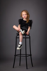 Little cute girl in black leather dress and sneakers posing on bar stool in studio on dark background