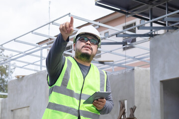 Fototapeta premium Asian overweight construction project foreman wearing white hard hat and reflective safety vest stands inspecting a construction project while holding a tablet and building plan in his hand.