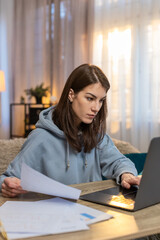 Young Caucasian woman sitting at table with laptop working on business plan looking focused typing text with documents spread around. Confident girl freelancer working from home in apartment room.