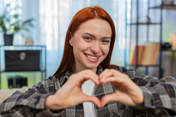 Young woman at home making heart shape with fingers smiling warmly to show love and affection....