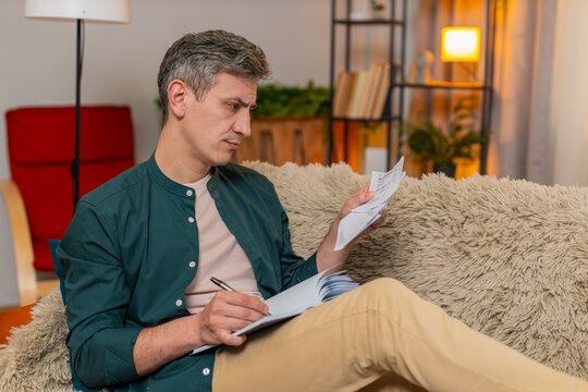 Young man at home seriously writes numbers in notebook while checking bills planning budget paperwork. Gray-haired guy on sofa stays focused and calm, managing household expenses with full attention - Powered by Adobe