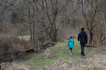 Fototapeta premium Strolling through a quiet forest path in early spring, a child and an adult enjoy a peaceful moment next to the gentle stream with trees emerging from winter's grasp