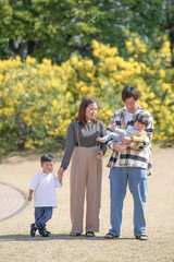Fototapeta premium In March, at a sunny forested park in Japan, a Japanese couple with their 3-year-old son and 6-month-old baby stands by a yellow flower field. This photo captures a quiet and loving family moment.