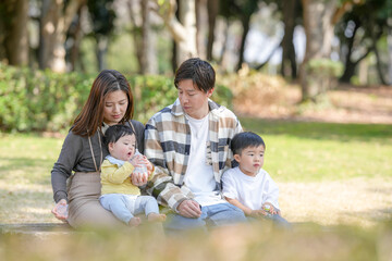 Fototapeta premium In March, at a sunny park in Fukuoka, a Japanese couple in their thirties sits on a bench with their 3-year-old son and baby girl, 6 months old. This photo captures a peaceful family moment outdoors.