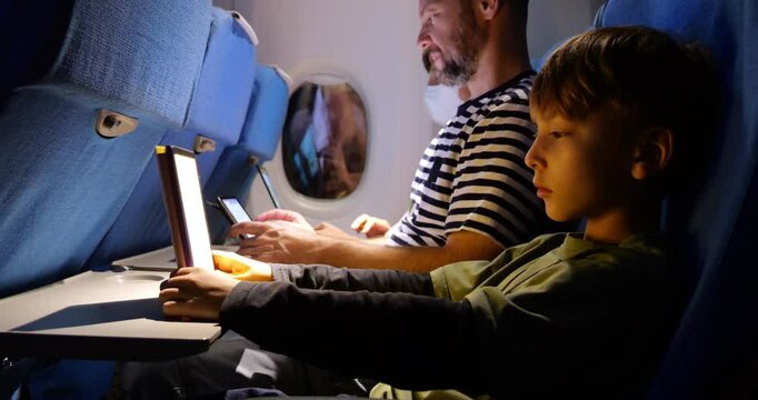 Nighttime airplane interior showing family during flight. Young boy reading on tablet or e-book device, while father uses tablet and mother works on laptop in background
