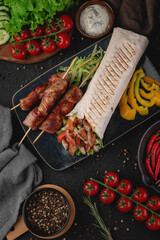 Close-up of grilled pork skewers served with a stuffed shawarma wrap, fresh vegetables like cucumber and tomato, and yellow bell pepper slices. Perfect for restaurant menus and ads