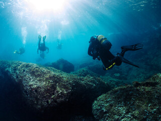 group of divers diving near the rocky bottom of the sea with sun rays shining from the surface,...