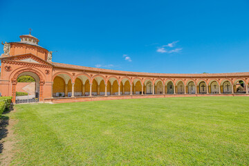 in Ferrara city of Italy, the ancient cemetery cloister featuring archway, tombs, and sculptures in the 15th-century Charterhouse of Ferrara, set against a lush green lawn with San Cristoforo church