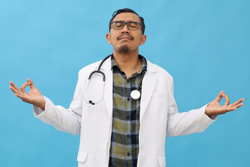 Calm doctor relaxed in meditation, breathing, stress free unaided at work, concept of conscious and peaceful adult male paramedic practicing yoga breathing exercises on blue wall.