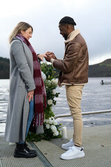 Smiling man putting engagement ring on woman's finger by lake