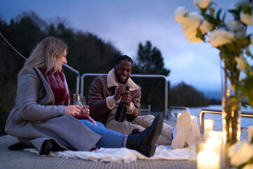 Smiling couple in warm clothing enjoying champagne on decorated pier at dusk