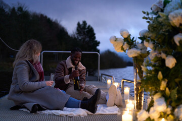 Smiling couple in warm clothing enjoying champagne on decorated pier at dusk