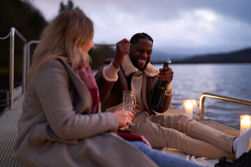 Smiling couple in warm clothing enjoying champagne on decorated pier at dusk