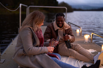 Smiling couple in warm clothing enjoying champagne on decorated pier at dusk