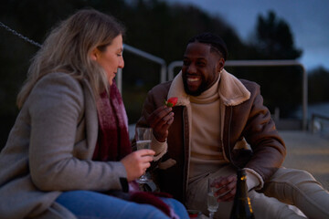 Smiling couple in warm clothing enjoying champagne and strawberries on pier